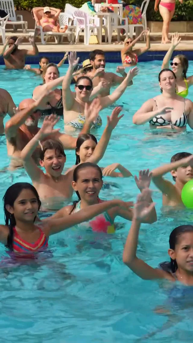 Piscina do clube cheia de crianças e adultos dançando de braços para cima